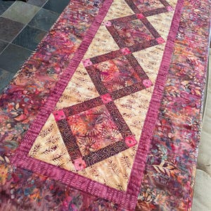 May include: A quilted table runner with a floral and geometric design. The runner features a central band of diamond-shaped blocks in shades of pink and burgundy, bordered by a patterned fabric with leaves and flowers. The overall color palette includes pinks, purples, and beige.