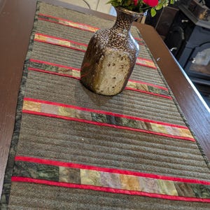 May include: A brown and green quilted table runner with red accents. The runner features horizontal stripes of patterned fabric and a dark green border. A speckled brown vase holds red flowers and greenery.