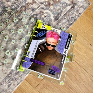 Pode incluir: Um fichário de plástico transparente com estampa de margaridas e um mecanismo de encadernação de argolas verde. O fichário tem uma página de revista dentro com uma foto de uma pessoa com cabelo rosa e o texto "ATEEZ THE WORLD EP. 2: OUTLAW" na página.