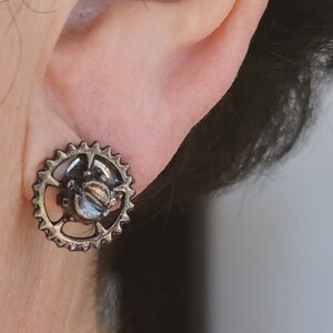 Peut inclure: Une paire de boucles d'oreilles en argent avec un motif d'engrenage. Les boucles d'oreilles sont portées à l'oreille d'une personne.