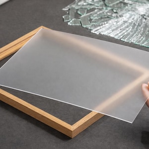 Könnte beinhalten: Ein Holzbilderrahmen mit einer Milchglasscheibe. Der Rahmen hat eine hellbraune Farbe und das Glas wird von einer Hand gehalten. Zerbrochenes Glas befindet sich im Hintergrund.