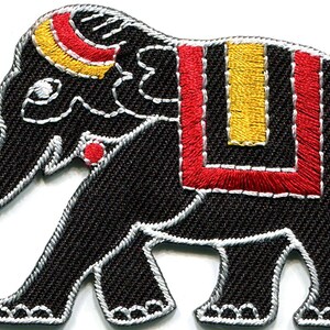 May include: An embroidered patch in the shape of an elephant. The elephant is black with silver thread outlining its body. It features a red and yellow headpiece, a red and yellow saddle blanket, and a red tail tip with a small red dot.