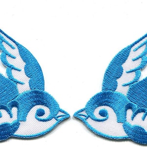 May include: Two embroidered swallow patches featuring a vibrant blue and white color scheme. Each bird is depicted in flight with wings spread, showcasing detailed stitching and a distinct silhouette facing toward the center of the frame.