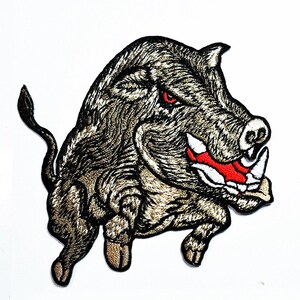 May include: An embroidered patch featuring a charging wild boar in shades of brown, tan, and black. The boar has a fierce expression with a bright red eye, a wide-open mouth showing white tusks, and a red tongue, all outlined in black thread.