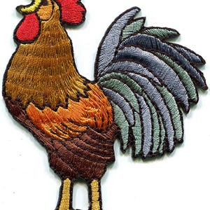 May include: An embroidered iron-on patch featuring a rooster with a bright red comb and wattle, a golden-brown neck and body, and a tail with layered feathers in shades of light blue, gray, and dark brown, standing on yellow legs.