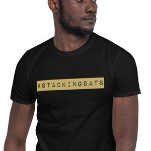 Puede incluir: Una camiseta negra con un rectángulo dorado que dice "#STACKINGSATS" en texto negro.