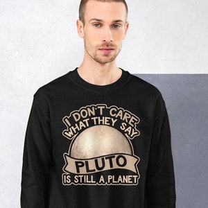以下が含まれることがあります： 「私は気にしない、彼らは言う、冥王星はまだ惑星だ」というテキストと、惑星冥王星のグラフィックプリントが施された黒いスウェットシャツ。
