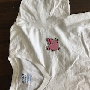 Puede incluir: Camiseta blanca de cuello en V con un cerdo de dibujos animados rosa bordado en la parte delantera. La camiseta tiene una etiqueta de marca Gildan en la parte inferior.