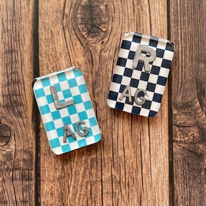 May include: Two small, rectangular, checkered, resin tiles with silver lettering. The tile on the left is blue and white with the letters "L AG". The tile on the right is black and white with the letters "R AG".