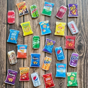 May include: A collection of miniature snack food and beverage replicas, including bags of chips like Doritos, Cheetos, and Lay's, and soda cans such as Coca-Cola, Pepsi, and Fanta, arranged on a wooden surface.
