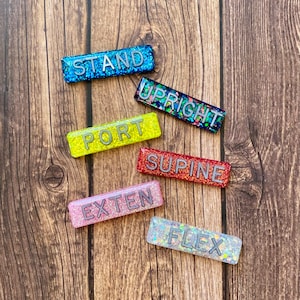 May include: Six rectangular resin plaques with glitter finishes in blue, dark multi-color, yellow, red, pink, and clear. Each plaque features raised grey lettering spelling out the words STAND, UPRIGHT, PORT, SUPINE, EXTEN, and FLEX respectively.