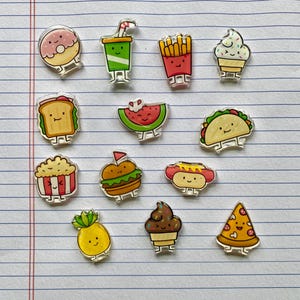 Op de afbeelding: Een verzameling cartoon-speldjes met eten, waaronder een donut, frisdrank, friet, ijs, een sandwich, watermeloen, taco, popcorn, een hamburger, een hotdog, ananas, cupcake en pizza. Elke speld heeft een zilveren rand.
