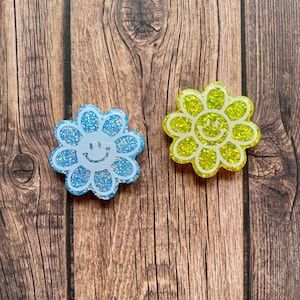 Puede incluir: Dos pines de flores con purpurina, uno azul y otro amarillo, con una carita sonriente blanca en el centro de cada flor.