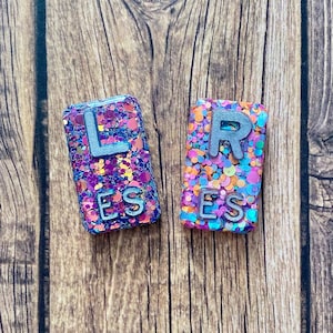 May include: Two rectangular, glitter-covered resin pieces. One has the letter "L" above "ES" in silver. The other has "R" above "ES" in silver. The glitter is a mix of pink, purple, blue, and orange. The background is a wooden surface.