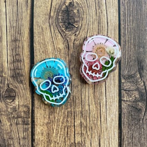 Puede incluir: Dos adornos de calavera de resina con contornos blancos, rellenos de flores prensadas. Una calavera está llena de flores rosas y blancas, la otra de flores azules y blancas.