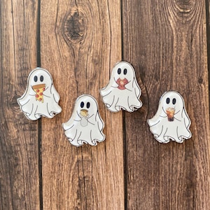 Puede incluir: Cuatro pines blancos con forma de fantasma con ojos negros y varios alimentos. Un fantasma sostiene pizza, otro un sándwich, un tercero una bebida y el último una taza de café. Los pines están sobre una superficie de madera marrón.