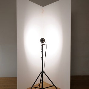 May include: A black photography light stand with a white reflector behind it. The light stand has a round light fixture attached to it.