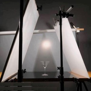 May include: A photography studio setup with two white backdrops, a wooden table, and a clear glass martini glass on the table.