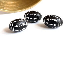 May include: Three black wooden beads with white geometric designs. The beads are oval shaped and have a glossy finish.