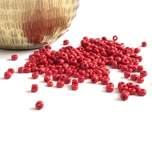 May include: A collection of small, red, round beads scattered on a white surface. The beads are a solid, matte color and appear to be made of glass or plastic. A gold-colored, textured bowl is partially visible in the background.