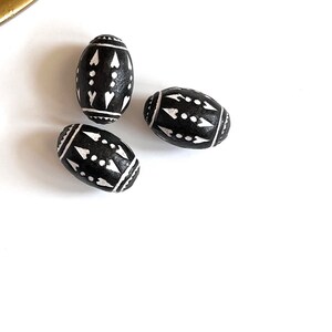 May include: Three black wooden beads with white geometric patterns. The beads are oval shaped and have a glossy finish.