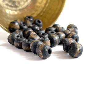 Puede incluir: Un montón de cuentas rayadas de color negro y dorado, que se derraman de un cuenco de latón.