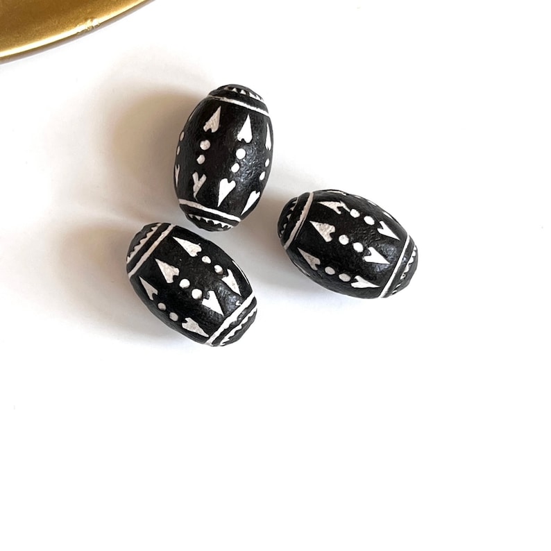 May include: Three black oval-shaped wooden beads with white geometric patterns. The beads have a black background with white geometric designs.