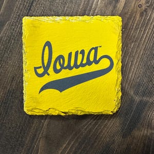 May include: Square yellow coaster with the word "Iowa" in a script font and a dark blue swoosh. The coaster has a rough, textured edge and is set against a dark wood grain background.