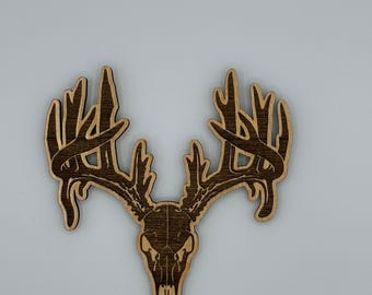 May include: Wooden deer skull with antlers, featuring a detailed design. The skull and antlers are a dark brown color, with a lighter brown outline. The antlers are intricately shaped, and the skull has detailed features.