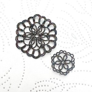 K&ouml;nnte beinhalten: Zwei schwarze und irisierende Blumenbroschen. Die gr&ouml;&szlig;ere Brosche hat einen Durchmesser von etwa 7,6 cm, die kleinere etwa 3,8 cm. Sie liegen auf einem wei&szlig;en, strukturierten Hintergrund mit kleinen, verstreuten Punkten.