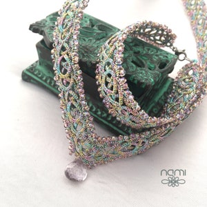 May include: A multi-colored beaded necklace with a delicate, intricate design. The necklace features a small, iridescent gemstone pendant and is displayed on a green box with the word "nami" written on it.