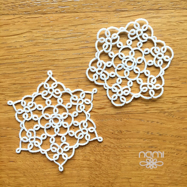 May include: Two white crocheted snowflakes on a wooden surface. The snowflakes are intricate and delicate, with a circular pattern.