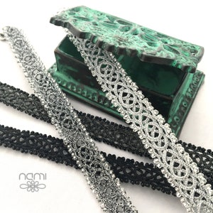 May include: Two spools of black and silver lace trim. The silver lace trim is in a decorative pattern and is on top of a green box. The black lace trim is underneath the silver lace trim. The word "nami" is printed on a white background.