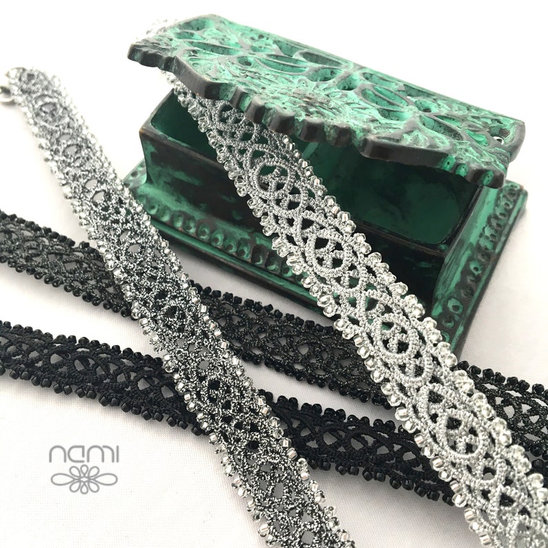 May include: Two spools of black and silver lace trim. The silver lace trim is in a decorative pattern and is on top of a green box. The black lace trim is underneath the silver lace trim. The word "nami" is printed on a white background.