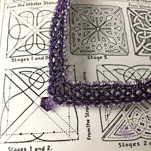 May include: A close-up of a purple beaded lace trim with a silver thread running through it. The trim is laid on top of a page from a book with black and white line drawings of geometric patterns. The text on the page reads "From the Ulbster Stone" and "Stages 1 and 2".