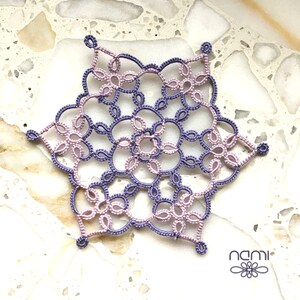 May include: A delicate purple and pink crocheted snowflake with a detailed design. The snowflake is made with fine thread and has a lacy appearance.