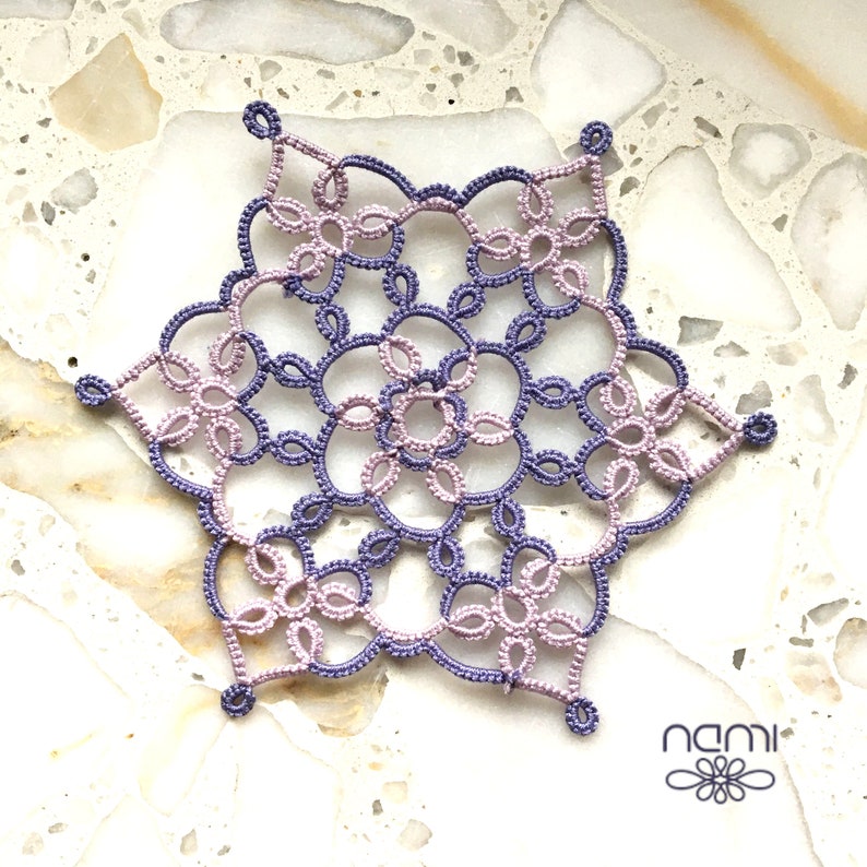 May include: A delicate purple and pink crocheted snowflake with a detailed design. The snowflake is made with fine thread and has a lacy appearance.