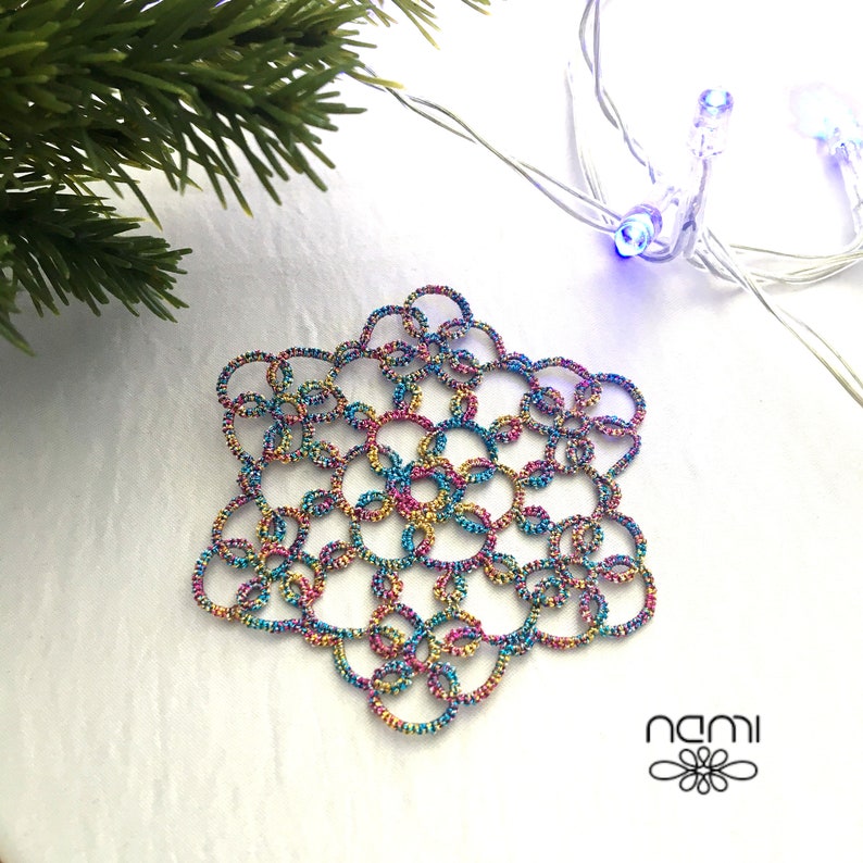 May include: A colorful snowflake ornament made of crocheted thread. The snowflake is a six-pointed star with a repeating pattern of loops and circles. The thread is a mix of pink, blue, green, and gold.