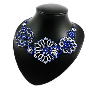 K&ouml;nnte beinhalten: Handgefertigte Halskette mit floralem Design in Wei&szlig; und Marineblau. Die Halskette wird auf einer schwarzen Schaufensterpuppe pr&auml;sentiert. Das filigrane, spitzenartige Muster erzeugt eine zarte und elegante &Auml;sthetik, ideal f&uuml;r besondere Anl&auml;sse.