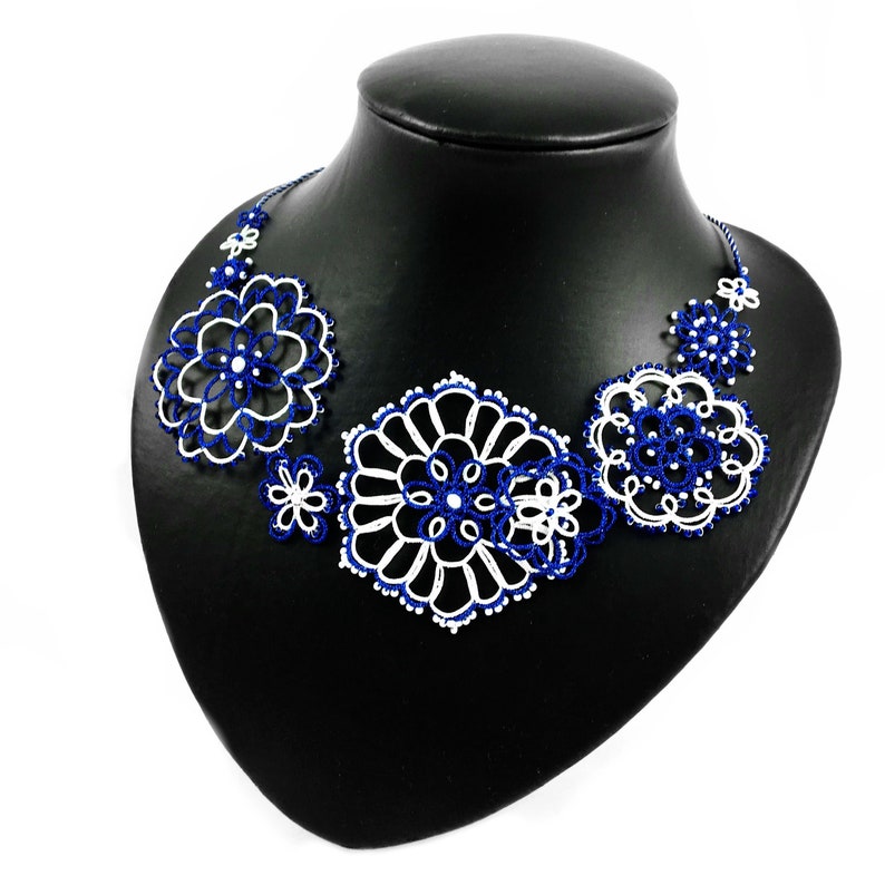 K&ouml;nnte beinhalten: Handgefertigte Halskette mit floralem Design in Wei&szlig; und Marineblau. Die Halskette wird auf einer schwarzen Schaufensterpuppe pr&auml;sentiert. Das filigrane, spitzenartige Muster erzeugt eine zarte und elegante &Auml;sthetik, ideal f&uuml;r besondere Anl&auml;sse.
