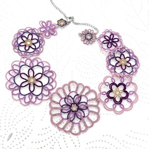 K&ouml;nnte beinhalten: Eine handgefertigte Halskette mit floralem Design. Die Halskette besteht aus lila und lavendelfarbenem Faden mit goldfarbenen Akzenten in der Mitte jeder Blume. Sie hat eine silberfarbene Kette und einen Verschluss.