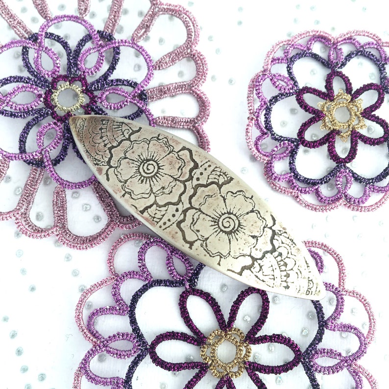 K&ouml;nnte beinhalten: Ein silberfarbenes, ovales Metallobjekt mit floralem Design liegt inmitten von drei handgefertigten Spitzendeckchen. Die Deckchen sind in Lila-, Rosa- und Goldt&ouml;nen gehalten und weisen komplizierte Muster auf. Der Hintergrund ist wei&szlig; mit kleinen silbernen Glitzerpunkten.