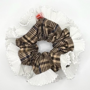 May include: A circular scrunchie featuring a brown plaid fabric at its centre, surrounded by a white pleated ruffle trim. The accessory is set against a white backdrop. A small, red tag is attached.