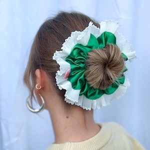 Könnte beinhalten: Haarstyling mit Dutt, verziert mit einem großen Haaraccessoire. Das Accessoire hat ein zentrales Element aus grünem Satin, umgeben von weißem, plissiertem Stoff. Silberne Creolen.