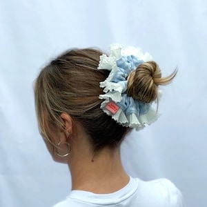 Puede incluir: Un coletero para el pelo con un centro de tela azul claro y un borde con volantes blancos. El accesorio se lleva alrededor de un moño e incluye una pequeña etiqueta rectangular de tela rosa con texto blanco en el lateral del coletero.