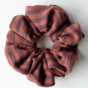 Può includere: Primo piano di un elastico per capelli con un motivo a righe. L'elastico è realizzato in un tessuto morbido e strutturato nei toni del ruggine e del marrone scuro. Le strisce corrono orizzontalmente sul tessuto, creando un design visivamente accattivante. È un accessorio per capelli.