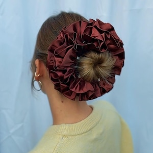 May include: A burgundy satin scrunchie with ruffled detailing, worn in a bun. The hair accessory is a deep red-brown colour and adds a touch of elegance. The scrunchie is a stylish hair accessory for various hairstyles.
