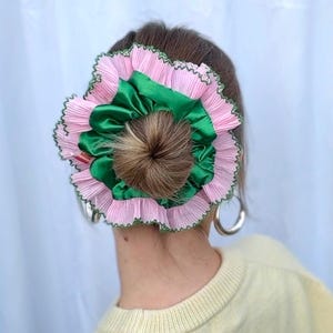 Könnte beinhalten: Ein rosa-grünes Haaraccessoire in Blumenform wird in einem Dutt getragen. Das Accessoire hat plissierten rosa Stoff mit grüner Bordüre und ein zentrales grünes Satinelement. Silberne Creolen sind sichtbar.