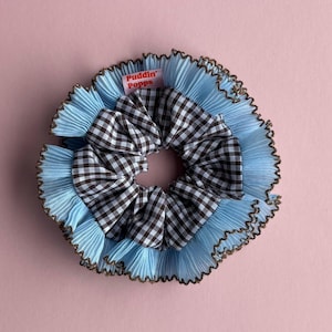 May include: A scrunchie with a light blue pleated fabric base, a black and white gingham center, and a brown trim. The scrunchie has a tag that reads "Puddin' Popps".