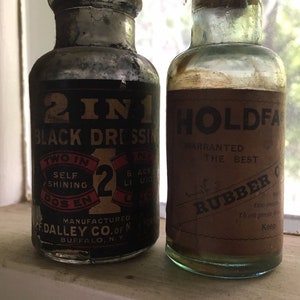 May include: Two glass bottles with labels. The bottle on the left is labeled "2 IN 1 BLACK DRESSING" and the bottle on the right is labeled "HOLDFAST WARRANTED THE BEST RUBBER OIL".